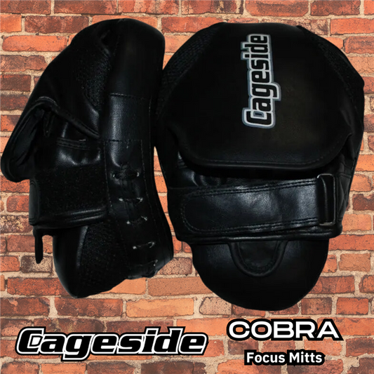 Black Cageside Cobra Focus Mitts against a brick wall background. These durable training mitts are designed with a secure velcro strap and ergonomic hand support for optimal control and precision during striking drills.