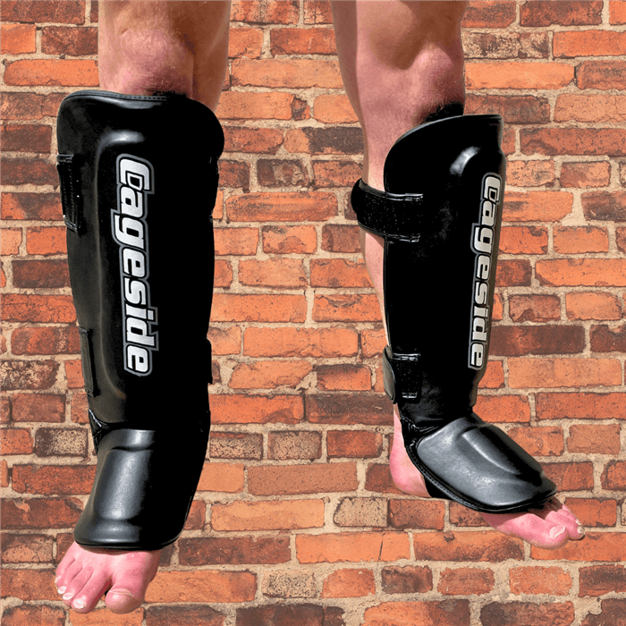 Front view of black Cageside Muay Thai shin guards worn on a person, featuring white vertical branding and adjustable hook-and-loop straps on a brick background.