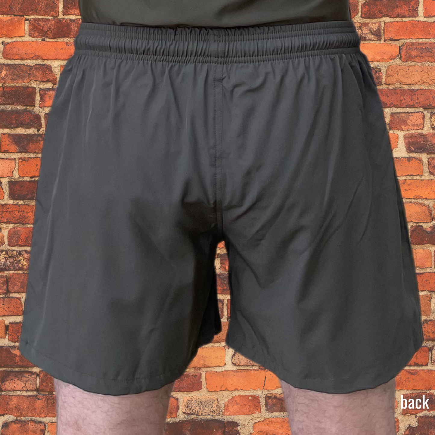 Back view of person wearing olive drab BJJ shorts with elastic waistband, displayed against a red brick background. The shorts provide a relaxed fit, ideal for training and casual wear.