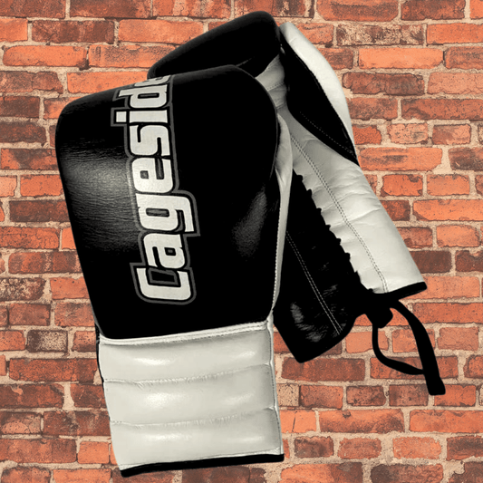 Pair of Cageside black and white lace-up boxing gloves hanging against a red brick wall, showing front and side padding.