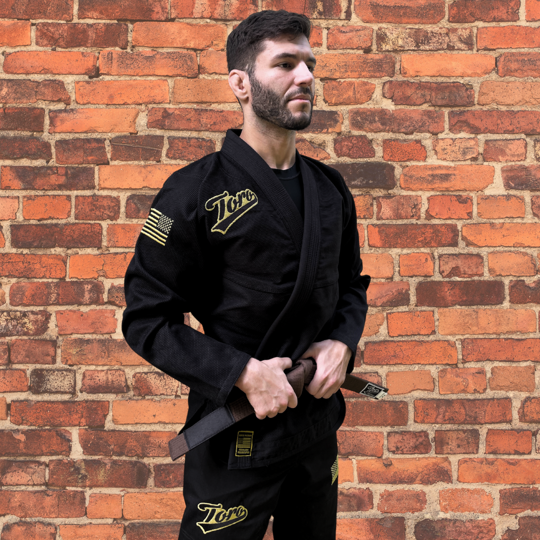 Male model in a black BJJ gi with gold Toro logo and USA flag patch, matching pants, and brown belt, standing in front of a brick wall.