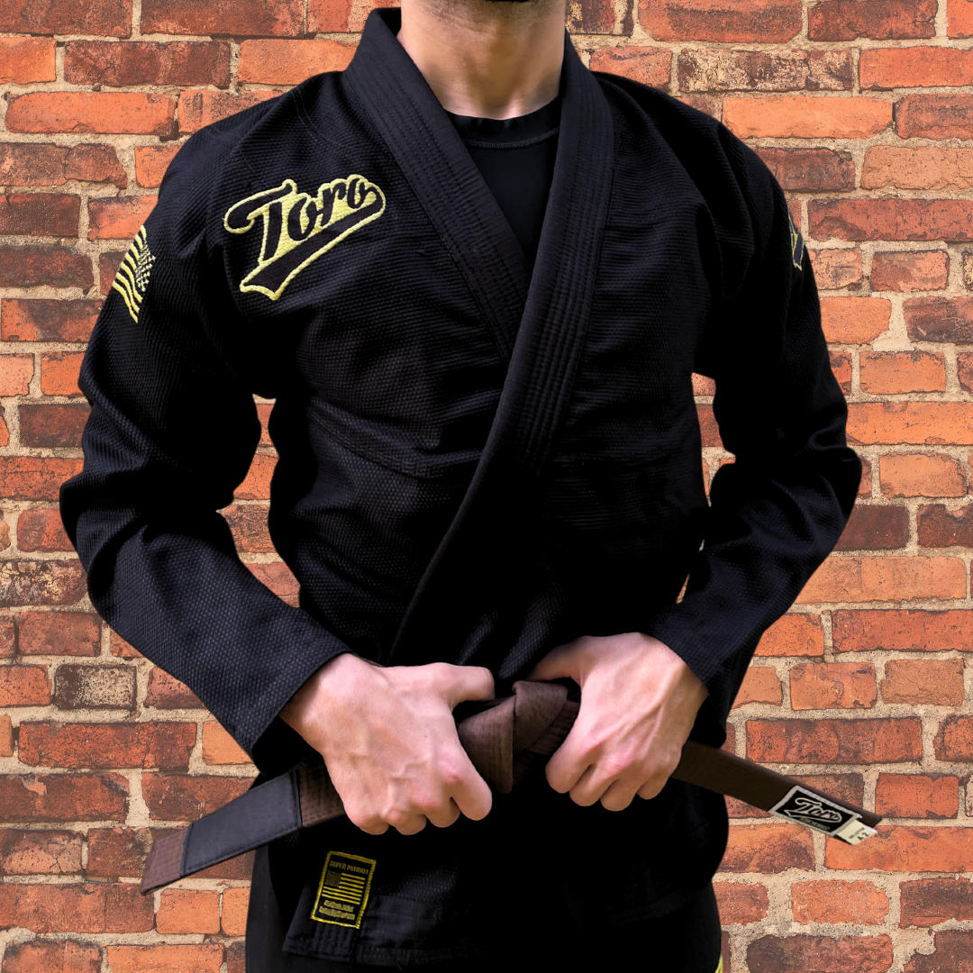 Black BJJ gi featuring a gold Toro logo and USA flag patch, shown on a male model wearing a brown belt, with a brick wall background.