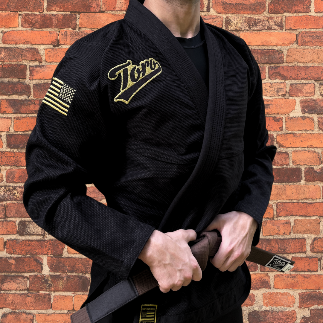 Black BJJ Gi with gold Toro logo and USA flag patch, competition legal, shown with a brown belt and brick wall backdrop.