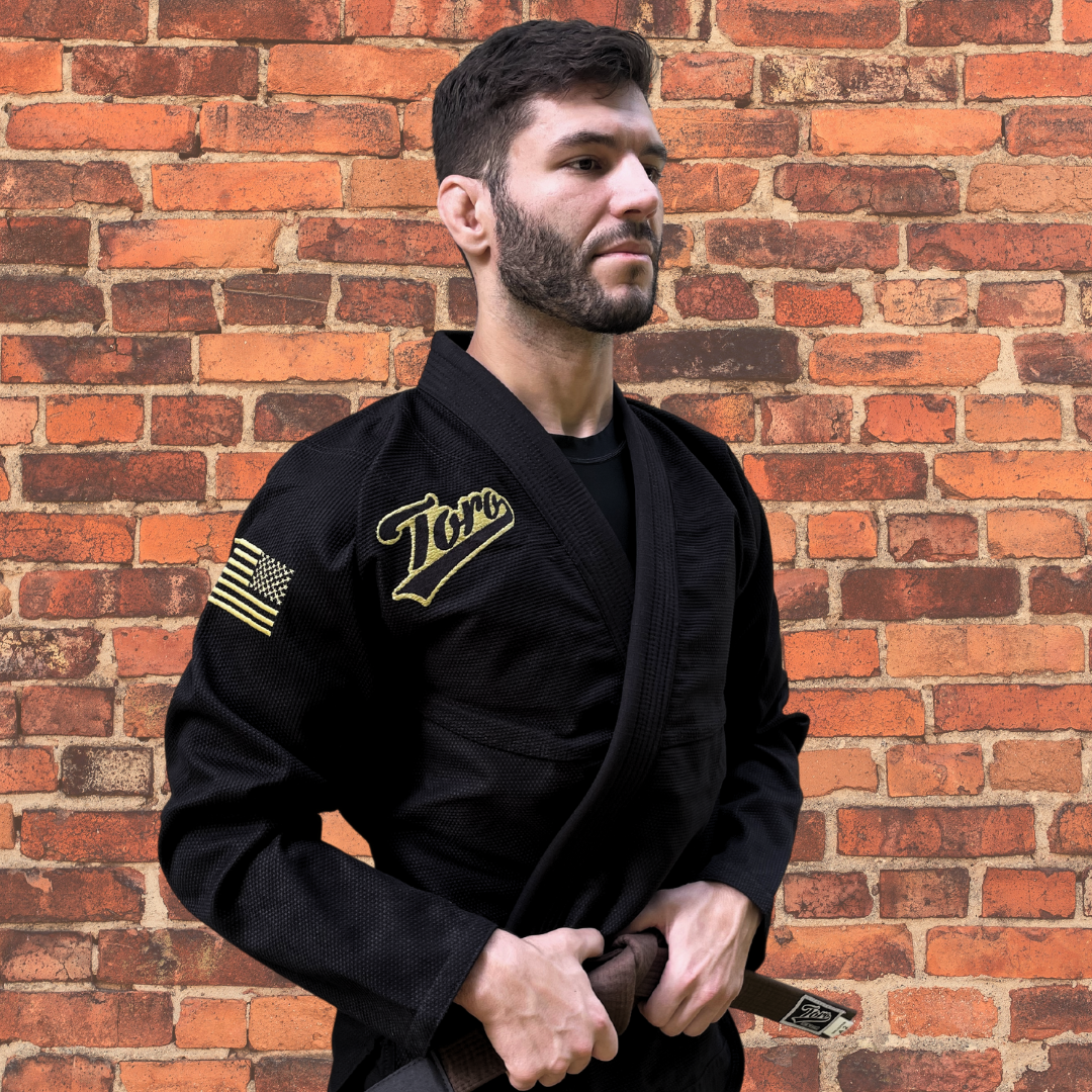 Black BJJ Gi with gold Toro logo and USA flag patch on sleeves, competition legal, brown belt, brick wall background.