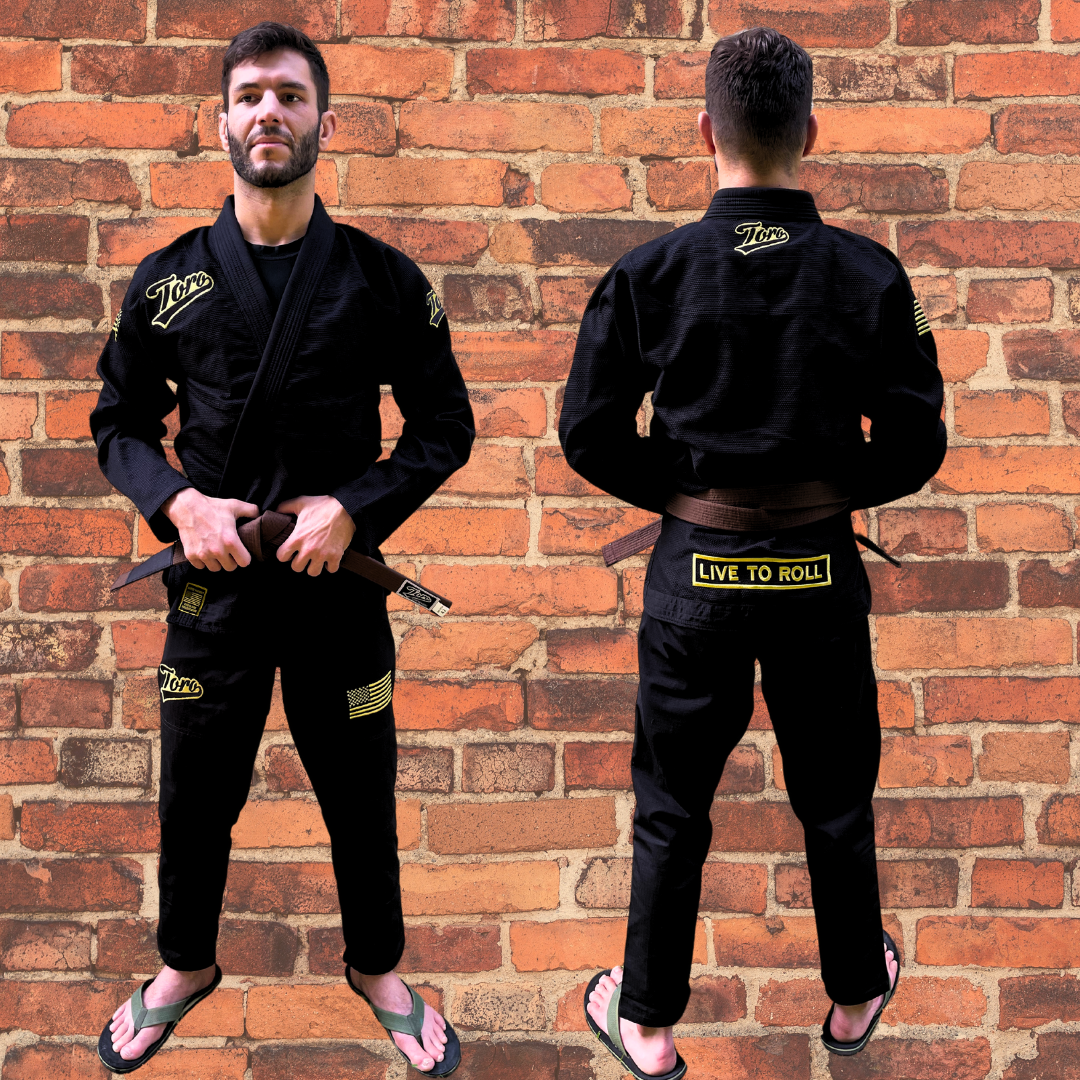 Male model wearing black BJJ gi with gold Toro logos, USA flag patch, and "Live to Roll" design on back, posing in front of brick wall.