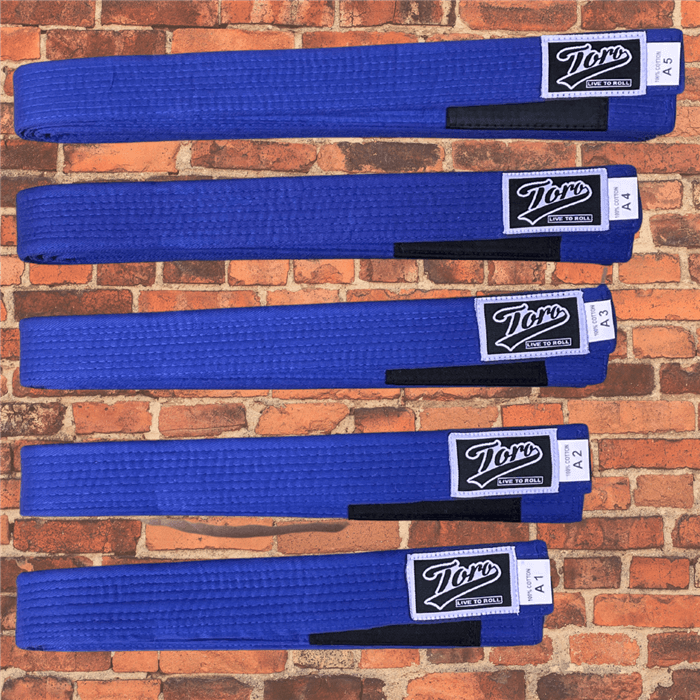 BJJ Blue belts by Toro, cotton construction, black stripe bar, sizes A1 to A5 displayed on brick wall background.