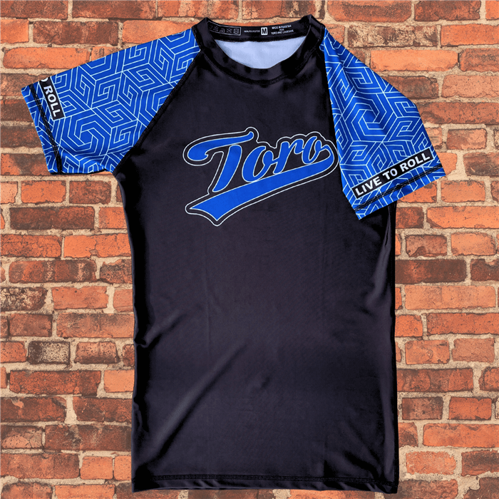 Flat lay of Toro BJJ Blue Belt Ranked Rash Guard with black body, blue geometric pattern sleeves, Toro logo on chest, Live to Roll text.