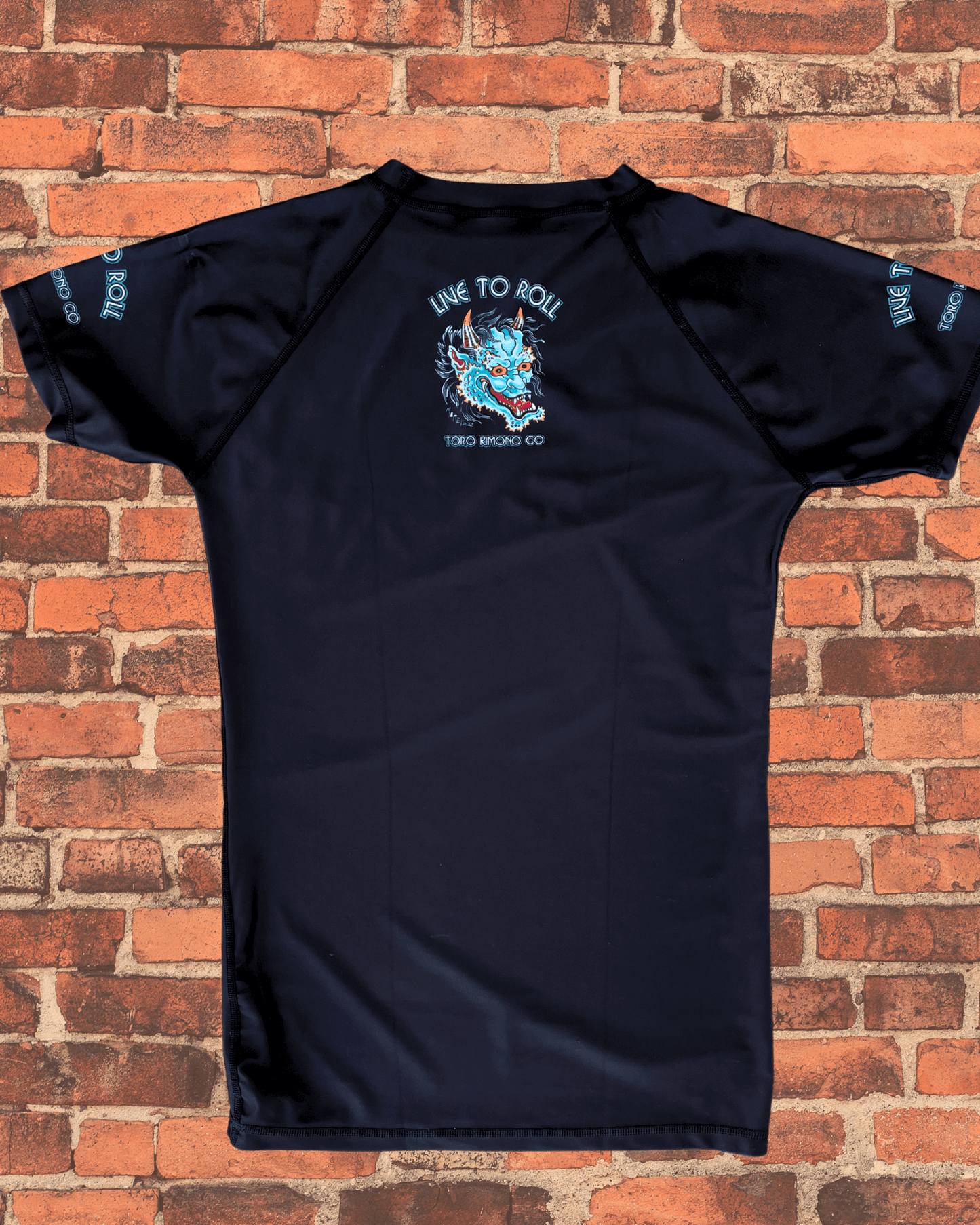 Back of the Toro BJJ Green Oni Rash Guard featuring a small demon mask design with “Live to Roll” and “Toro Kimono Co” text centered between the shoulder blades.