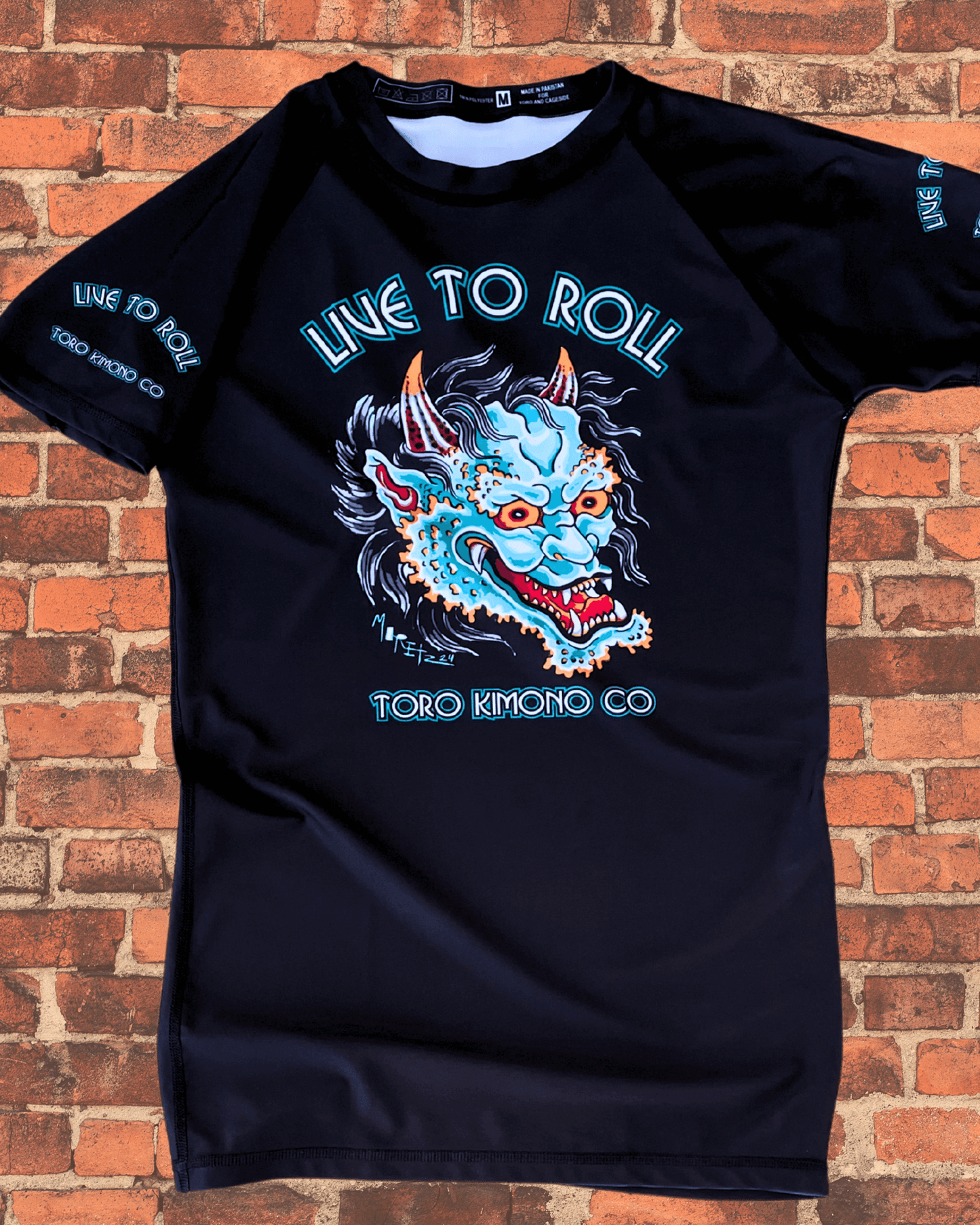 Front of the Toro BJJ Green Oni Rash Guard featuring a fierce blue oni demon mask with “Live to Roll” and “Toro Kimono Co” text on a dark background.
