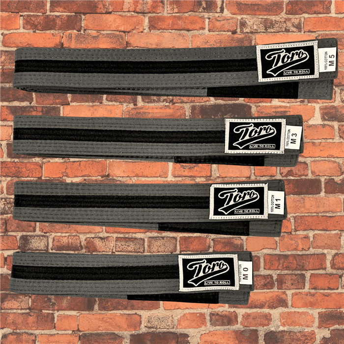 Toro BJJ kids gray and black BJJ belts in sizes M000–M5, featuring Toro logo patch with 'Live to Roll' text, black rank bar, brick background.
