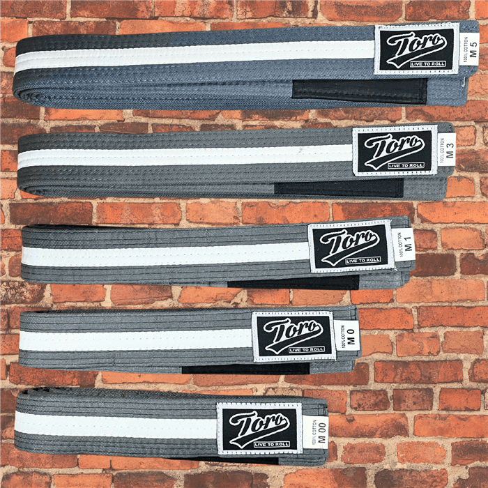 Toro BJJ kids grey belt with white stripe, displayed in sizes M000 - M5 on a brick wall background.