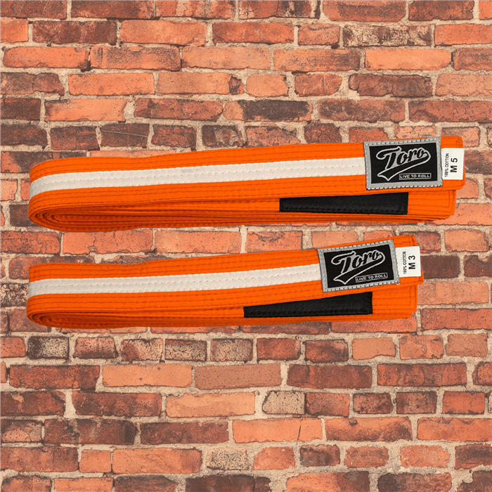 Toro BJJ kids orange BJJ belts with white stripe, sizes M000–M5, black rank bar, Toro logo patches, displayed on brick background.