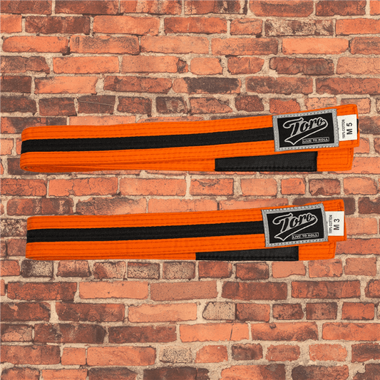 Toro BJJ kids orange and black BJJ belts in sizes M000–M5, featuring black rank bars and Toro logo patches, displayed on a brick background.