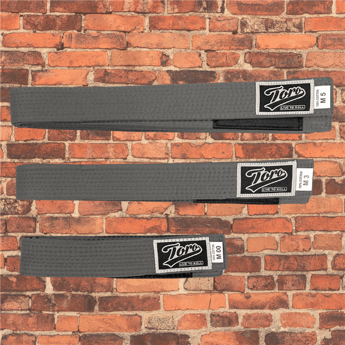 Toro BJJ kids' solid gray BJJ belts in sizes M000–M5, featuring a Toro logo patch with 'Live to Roll' text, a black rank bar, and a brick wall background.