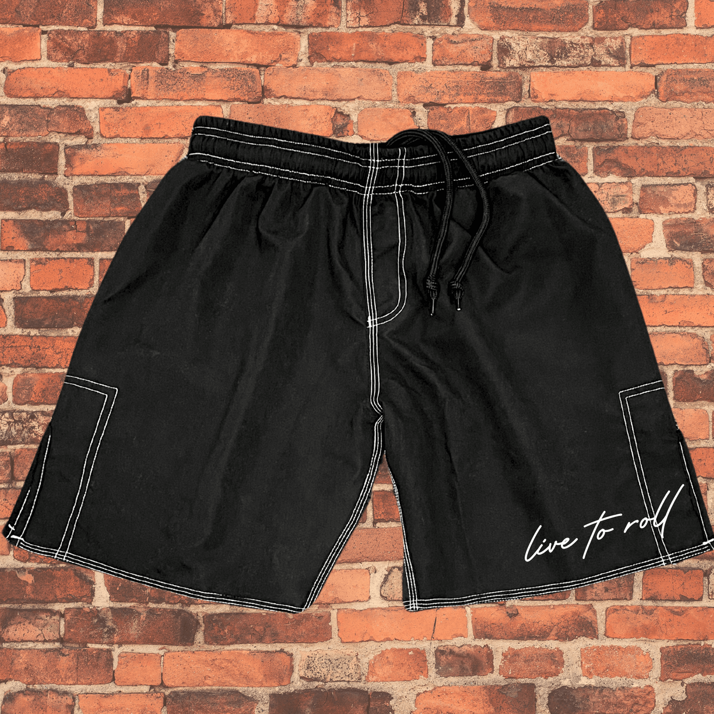 Flat lay of Toro BJJ Live to Roll black shorts with white stitching, side pocket, drawstring, for MMA, NoGi Jiu-Jitsu, and lounging.