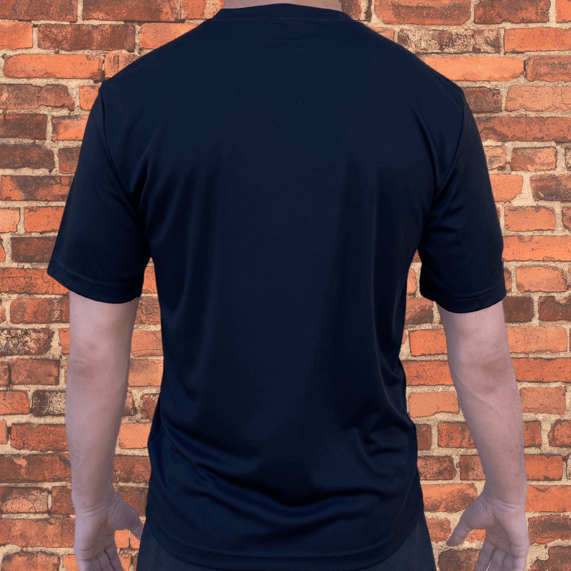 Back view of Toro BJJ Loose Fit Rash Guard in black, plain design, short sleeves, worn by male model, brick wall background.