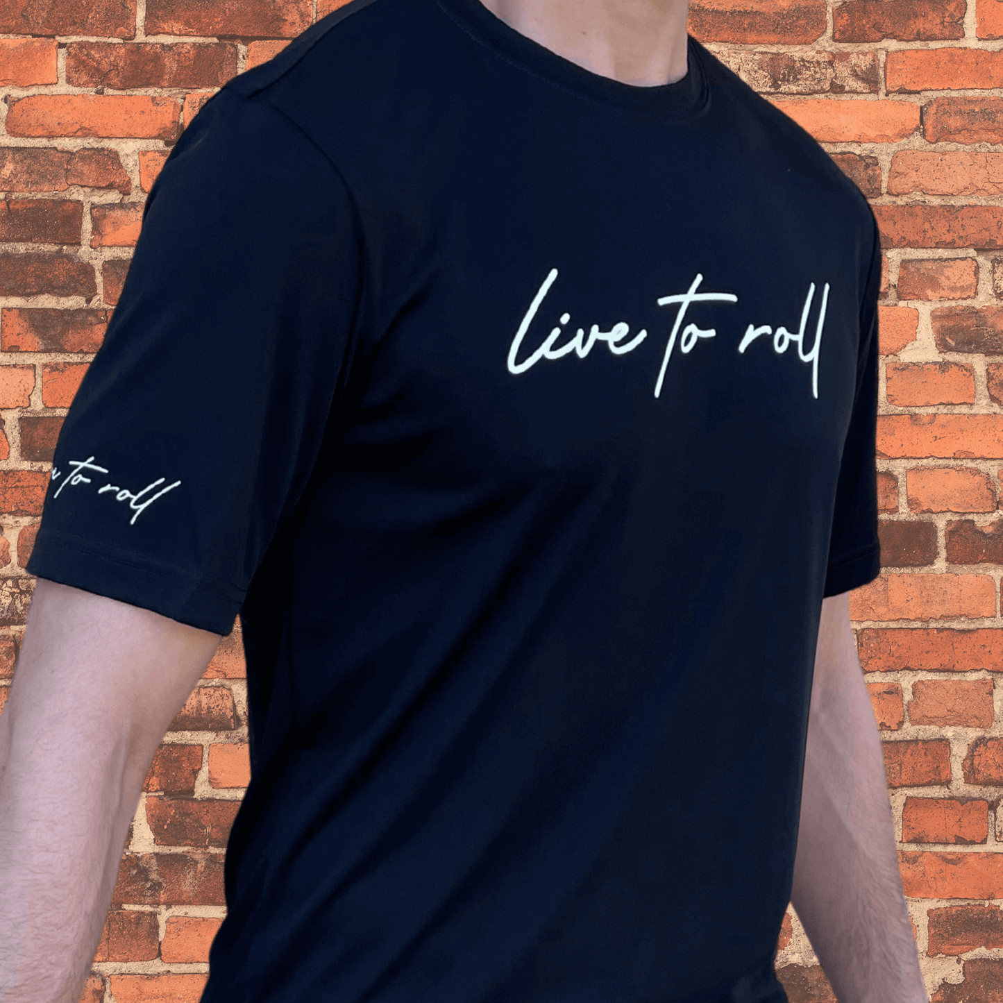 Side view of Toro BJJ Loose Fit Rash Guard in black, with "live to roll" text on chest and sleeve, worn by male model, brick wall background.