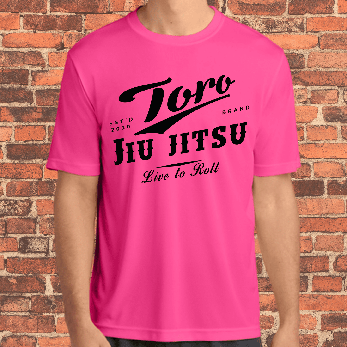 Adult-sized neon pink loose fit rash guard with bold black Toro Jiu Jitsu "Live to Roll" design, worn by a model in front of a brick wall.