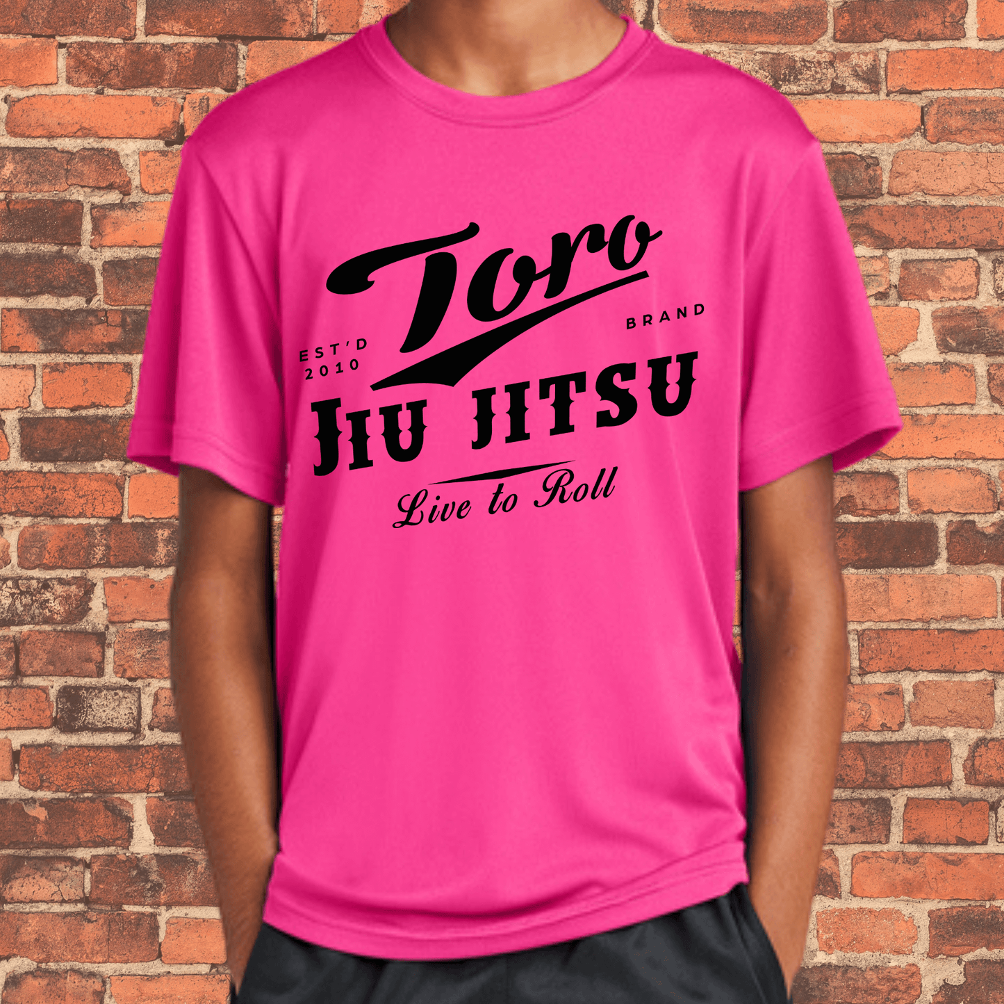 Kids-sized neon pink loose fit rash guard featuring Toro Jiu Jitsu "Live to Roll" design in bold black, worn by a young model in front of a brick wall.