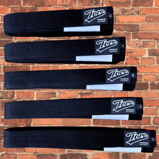 Premium black BJJ belt with white bar, Toro logo patch, and pearl weave material, designed for competition use, displayed on a brick wall background.