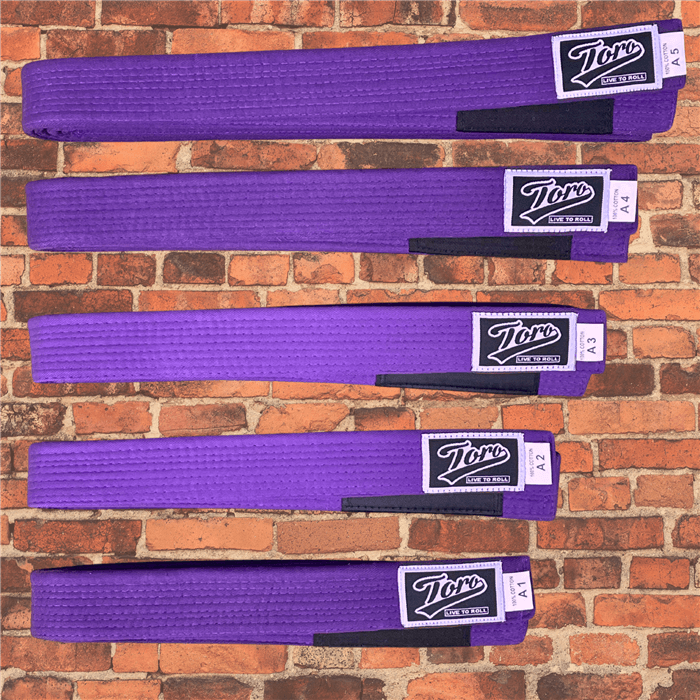 Purple BJJ belts by Toro, cotton construction, black stripe bar, sizes A1 to A5 displayed on brick wall background.