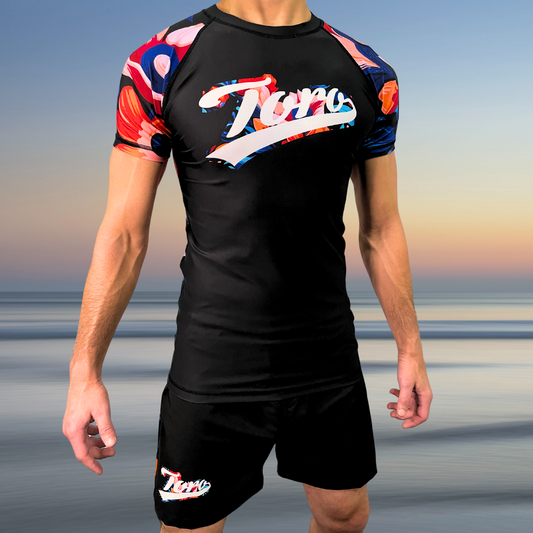 Toro BJJ NoGi kit with floral sleeve rash guard and black shorts with Toro logo, worn by male model, standing on beach background.