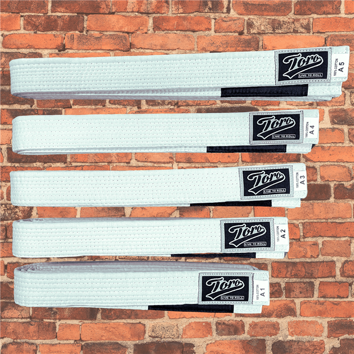 White BJJ belts by Toro, cotton construction, black stripe bar, sizes A1 to A5 displayed on brick wall background.