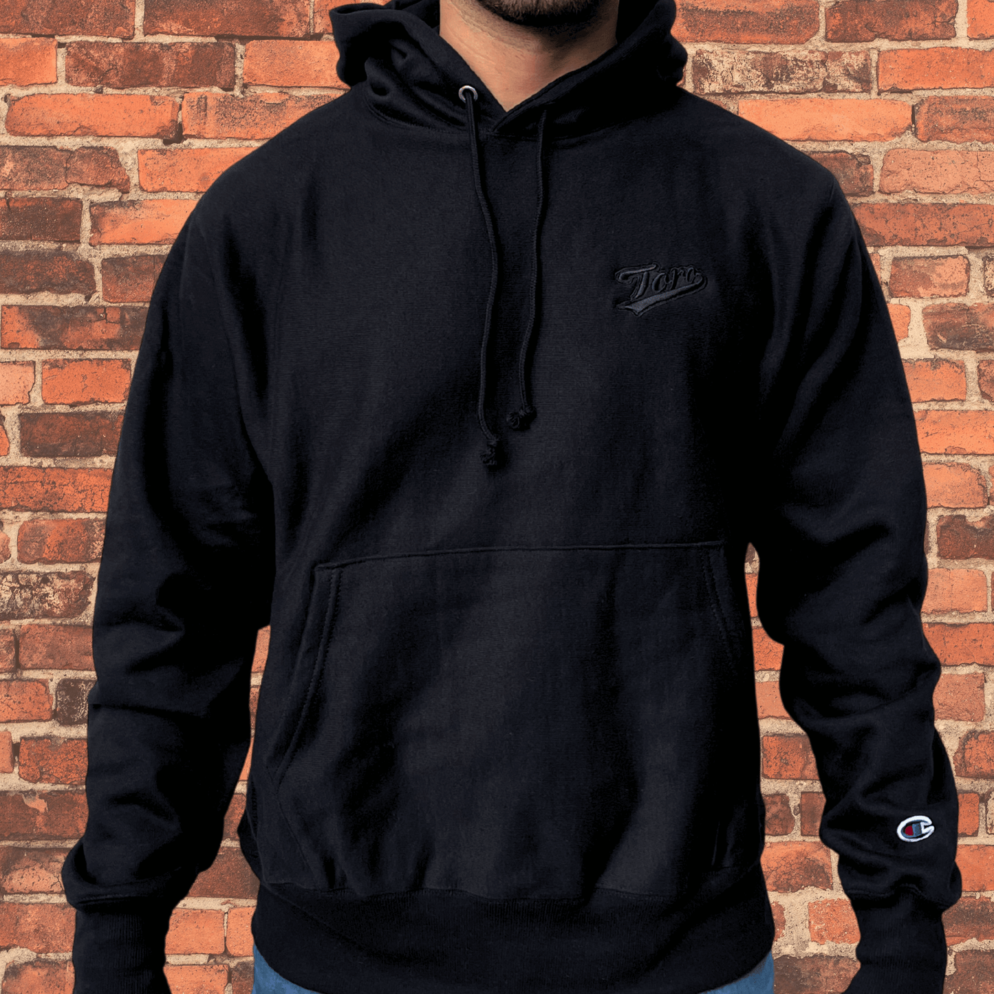 Model wearing Toro x Champion Reverse Weave Hoodie black on black with embroidered Toro logo and Champion logo on sleeve.