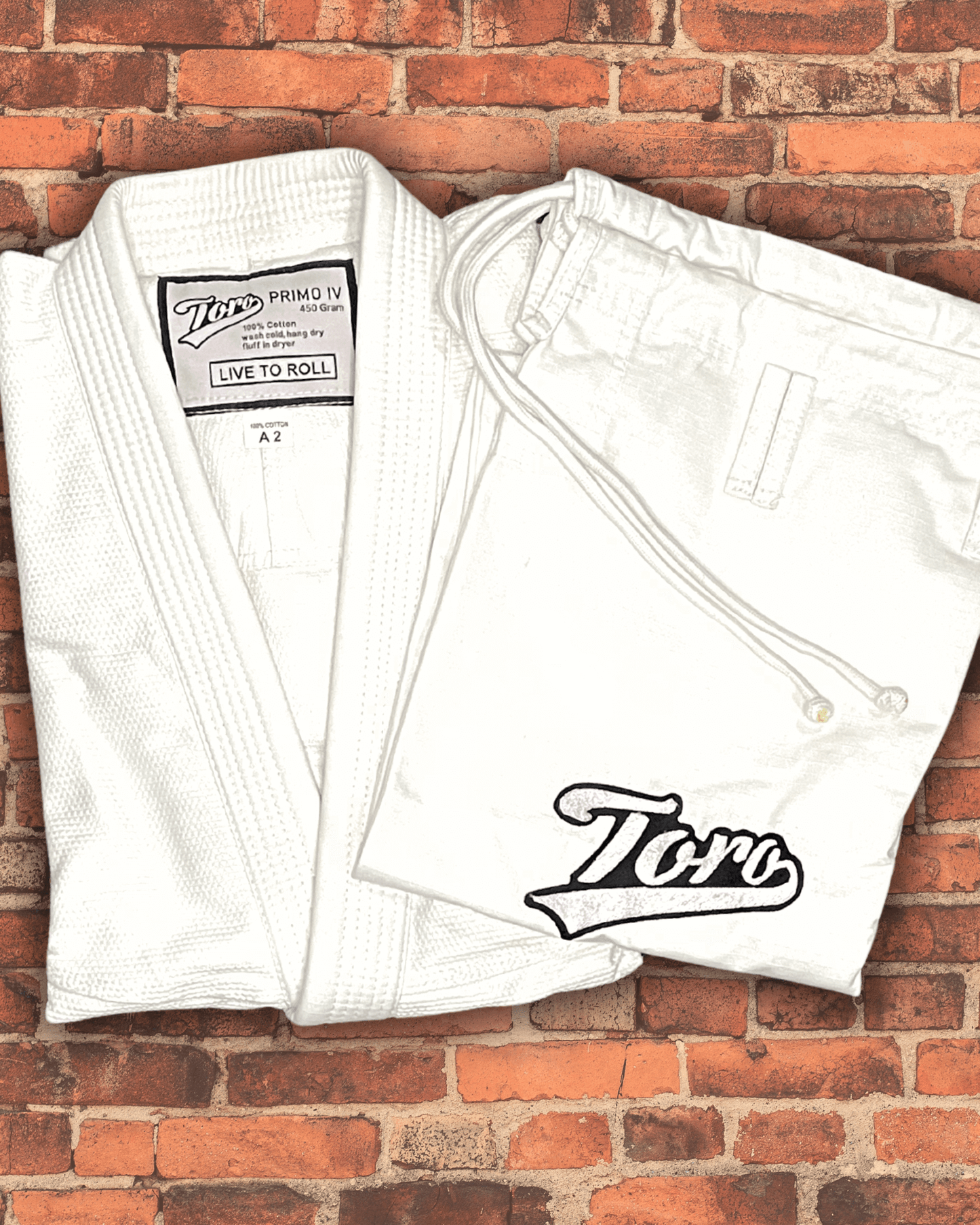 White Toro Primo BJJ Gi with 'Live to Roll' patch and 450-gram pearl weave fabric, folded on a brick background.