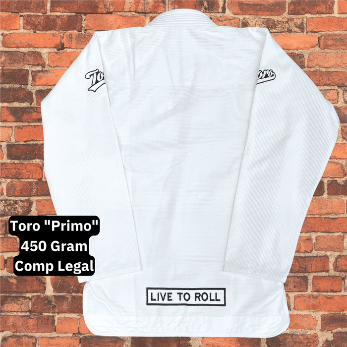 Back view of the Toro Primo White BJJ Gi jacket displayed flat against a brick wall background, featuring "Live to Roll" patch and shoulder logos with text highlighting 450 gram comp legal gi.