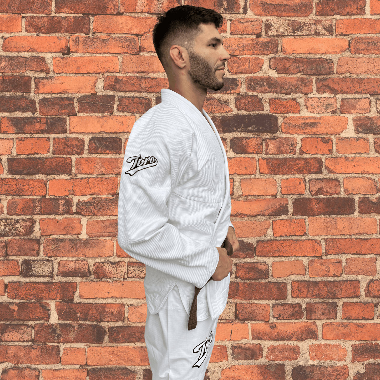 Male athlete wearing the Toro Primo white BJJ Gi with brown belt, standing against a brick wall. Overlay text highlights it as a 450 gram, comp-legal flagship gi ideal for training and competition.