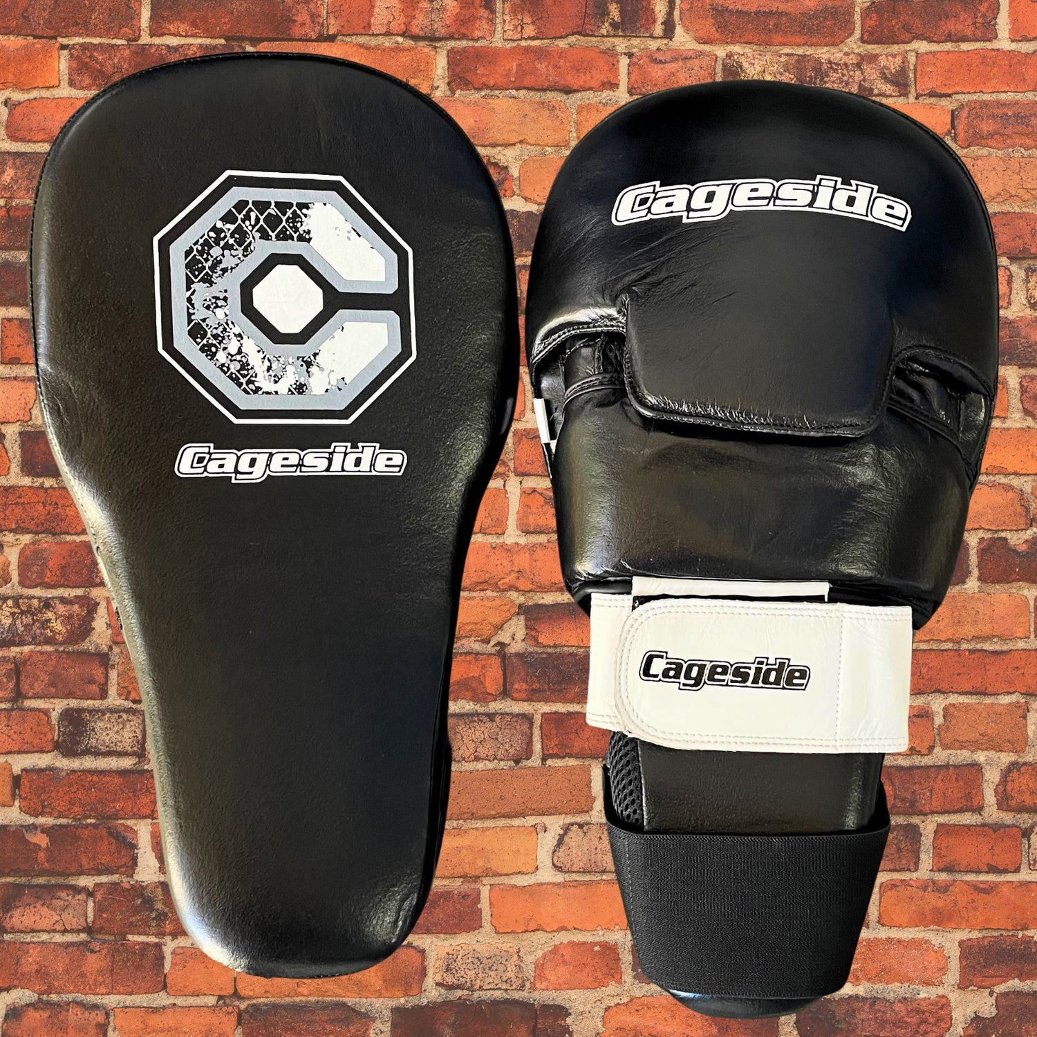 Black Cageside Cobra Focus Mitts against a brick wall background. These durable training mitts are designed with a secure velcro strap and ergonomic hand support for optimal control and precision during striking drills.