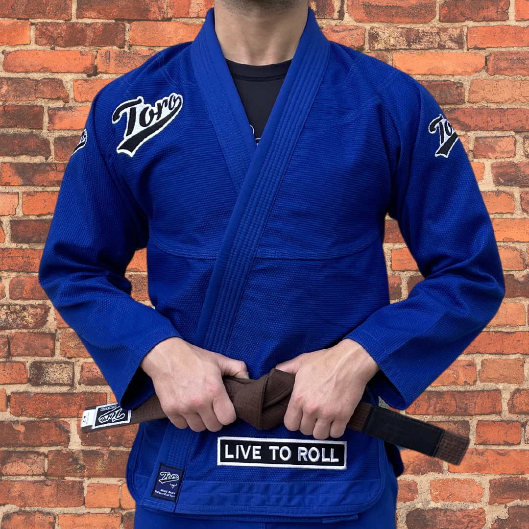 A BJJ athlete wearing a blue Toro Gi featuring the "Live to Roll" patch | Standing against a brick wall backdrop.