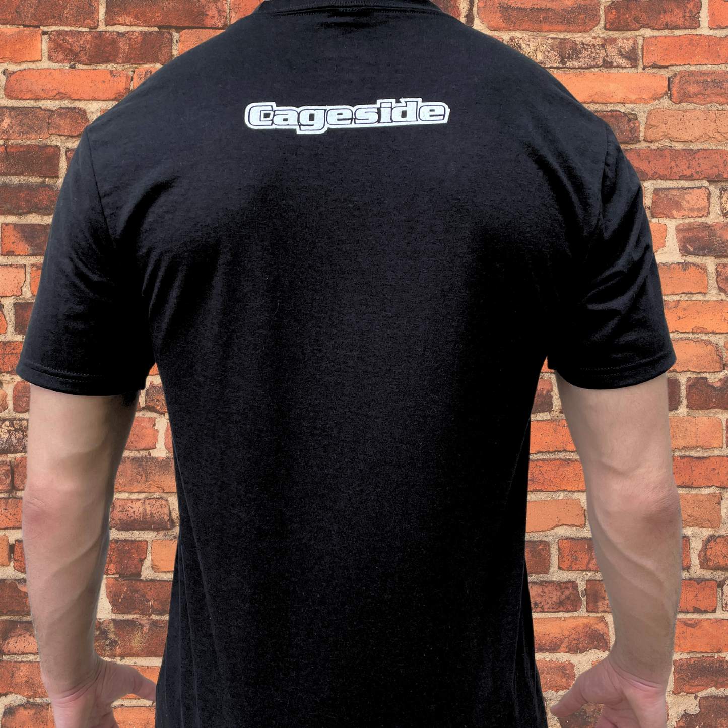 Back view of a black Muay Thai T-shirt displaying the Cageside logo centered across the upper back against a brick wall background.