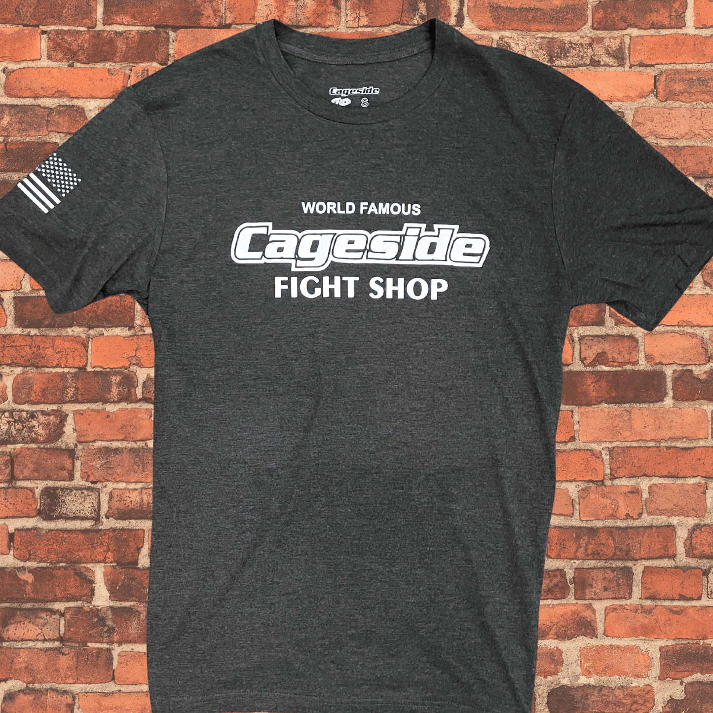 Flat lay view of charcoal Cageside Fight Shop t-shirt with white logo on brick background.