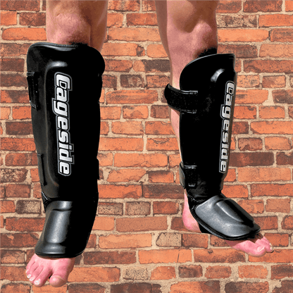 Front view of black Cageside Muay Thai shin guards worn on a person, featuring white vertical branding and adjustable hook-and-loop straps on a brick background.