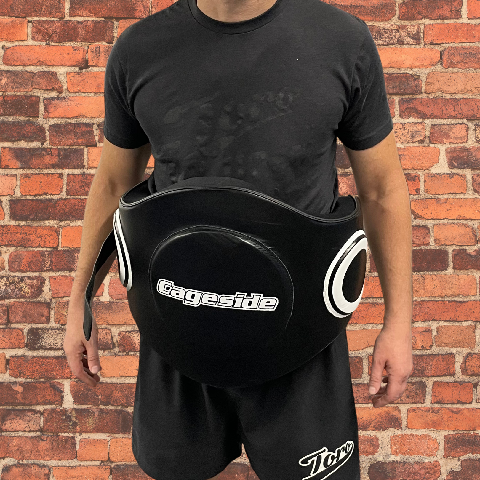 Person wearing Cageside's black protective body pad, ideal for MMA and striking training. The pad features side and center target zones for impact training, providing maximum protection while allowing freedom of movement. Displayed against a red brick wall, the gear ensures durability and performance.