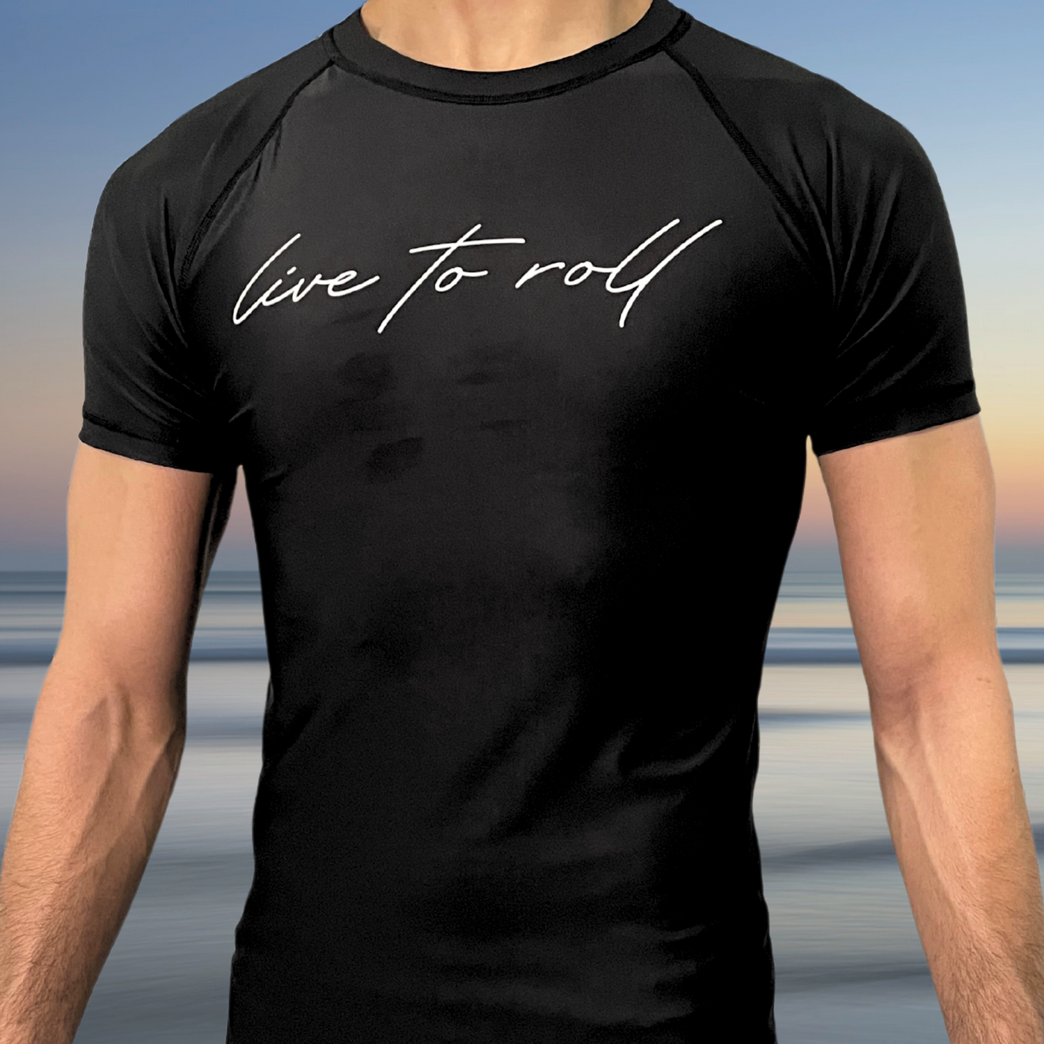 Black short-sleeve BJJ rash guard with “Live to Roll” script logo, worn by male athlete, beach background, Toro BJJ NoGi gear