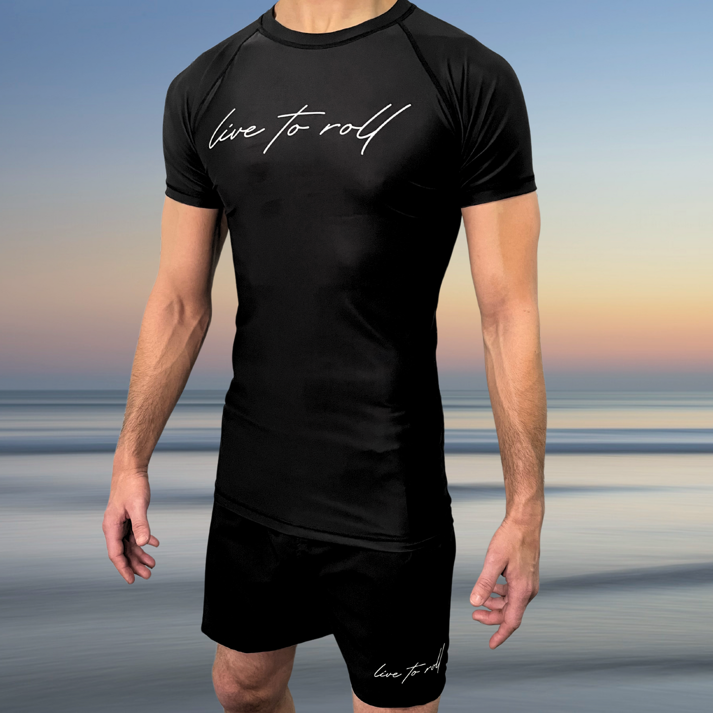 Black NoGi rash guard and shorts set with “Live to Roll” script, worn by male athlete standing on beach, Toro BJJ minimalist grappling gear