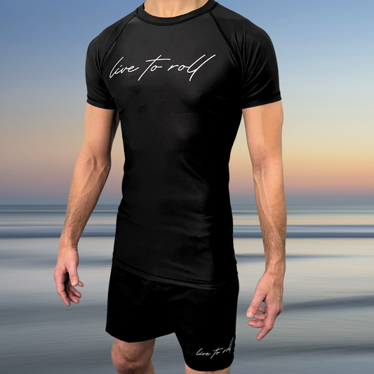 Black NoGi rash guard and shorts set with “Live to Roll” script, worn by male athlete standing on beach, Toro BJJ minimalist grappling gear