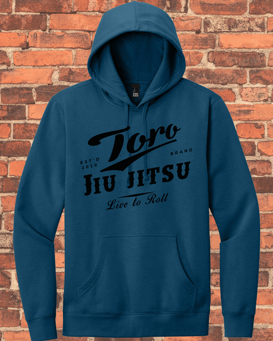 A neptune blue pullover hoodie with the "Toro Jiu Jitsu Est'd 2010 Brand Live to Roll" logo in black script and block lettering, displayed on a red brick background.