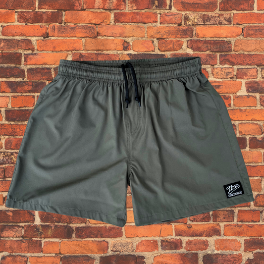 Olive drab BJJ shorts with elastic waistband and black drawstring, displayed against a red brick background. Features a small Toro BJJ patch on the lower right leg.