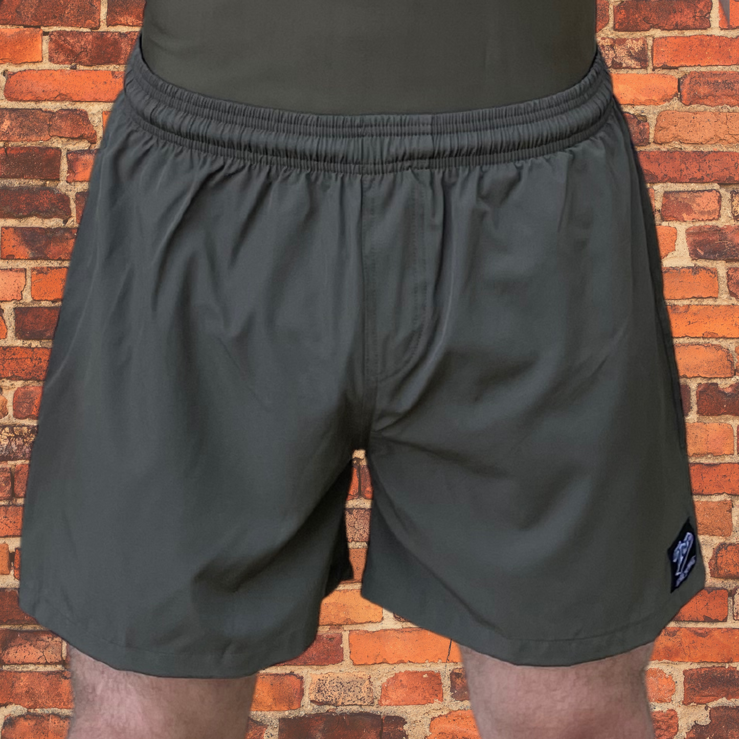 A person wearing olive drab BJJ shorts with elastic waistband and black drawstring, shown from the front against a red brick background. Features a small Toro BJJ patch on the lower right leg.
