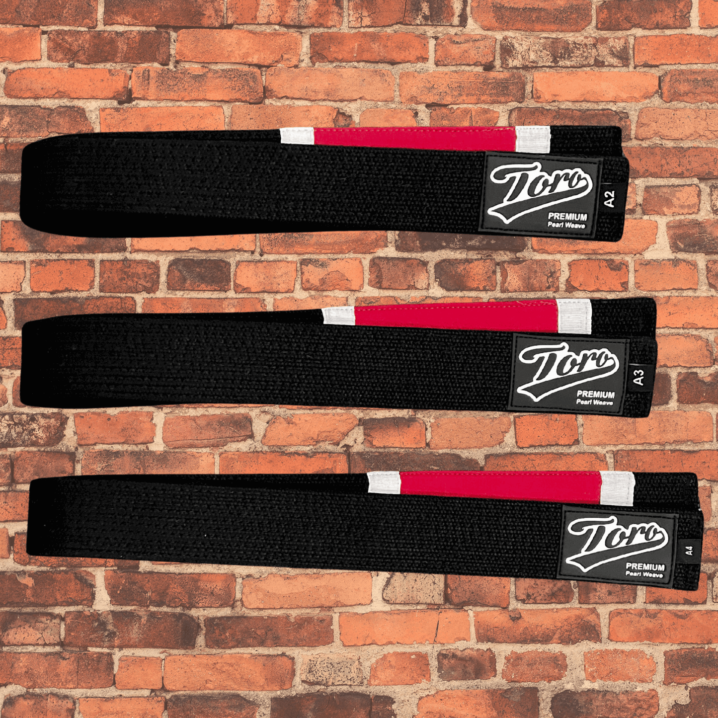 Toro Premium Pearl Weave Black BJJ Belts with professor stripes displayed on a brick wall background.