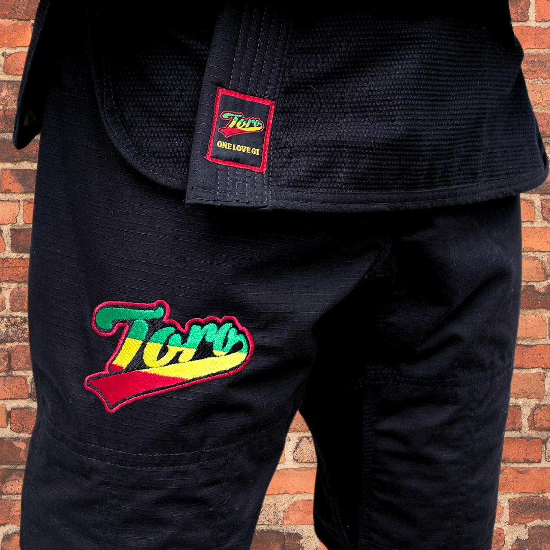 Close-up of Toro's black BJJ Gi with vibrant Rasta logo and 'One Love Gi' patch, set against a red brick wall backdrop.