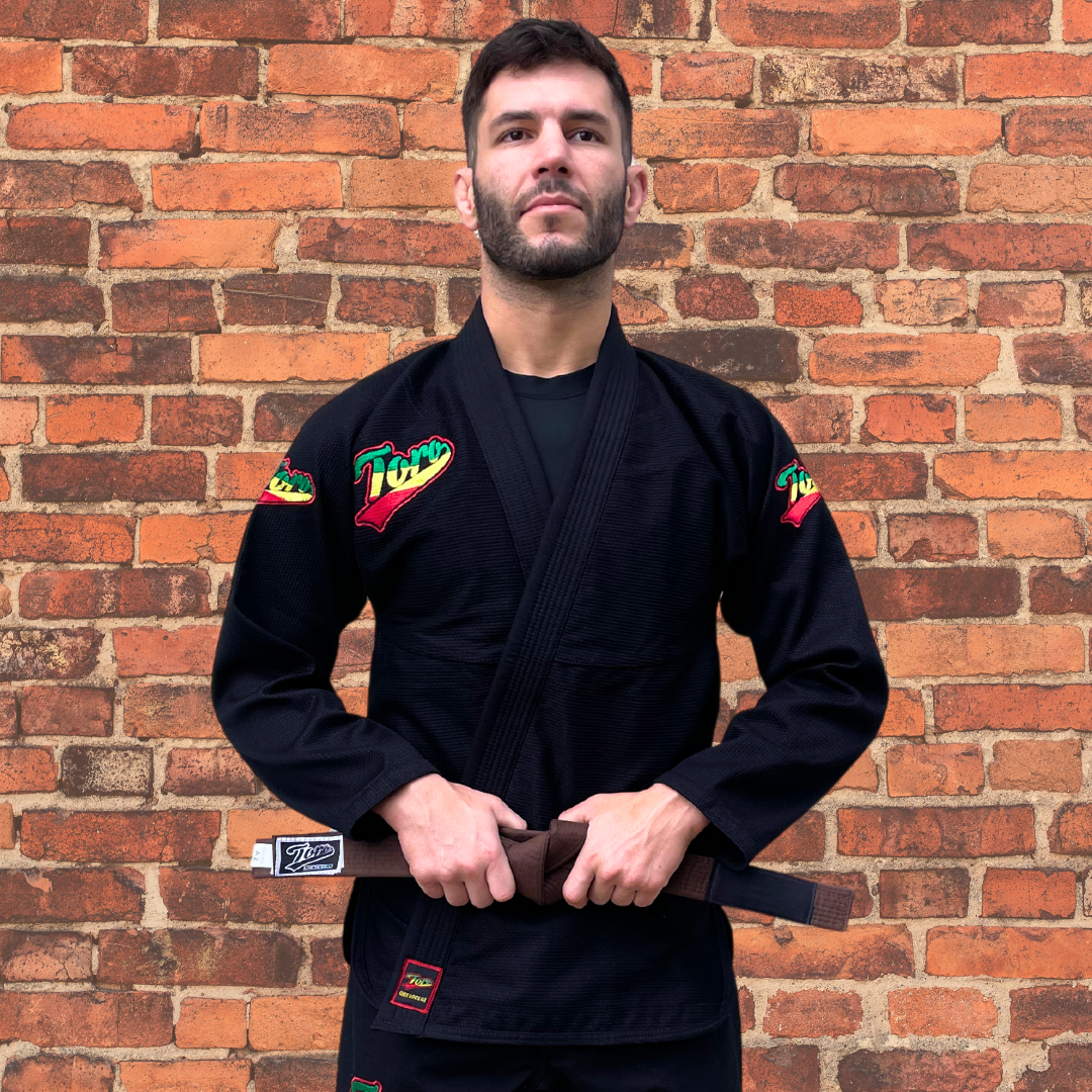 A man wearing Toro's black BJJ Gi with Rasta-themed logos and a brown belt poses against a red brick wall.