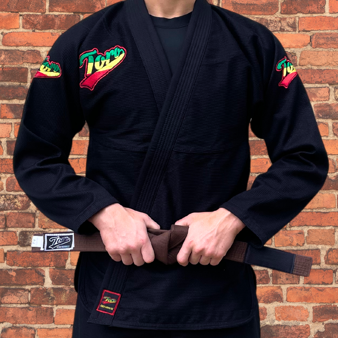 A man is wearing Toro's black BJJ Gi with Rasta-colored logos and a brown belt, perfect for Brazilian Jiu-Jitsu training.