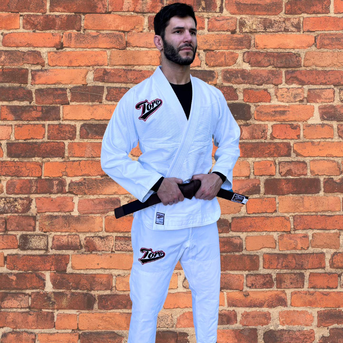 Man wearing Toro's white BJJ Gi with a brown belt, standing confidently against a red brick wall backdrop. The Gi features the Toro logo on both the jacket and pants, showcasing a clean, lightweight design perfect for competition or training. Ideal for Brazilian Jiu-Jitsu practitioners seeking durable and comfortable gear