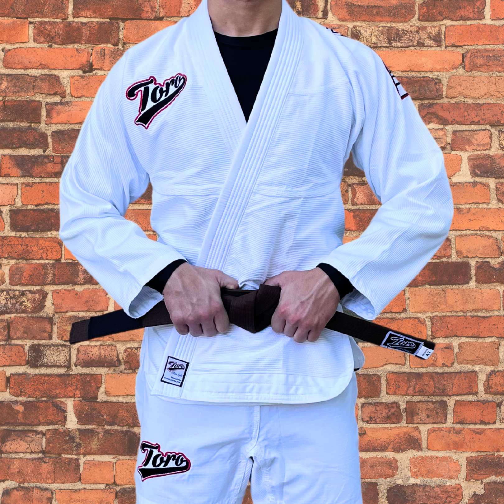 Brazilian Jiu-Jitsu practitioner wearing a white Toro BJJ gi with matching pants and a brown belt, standing in front of a brick wall. The lightweight gi features embroidered Toro logos on both the gi top and pants, offering a durable and comfortable option for training and competition.