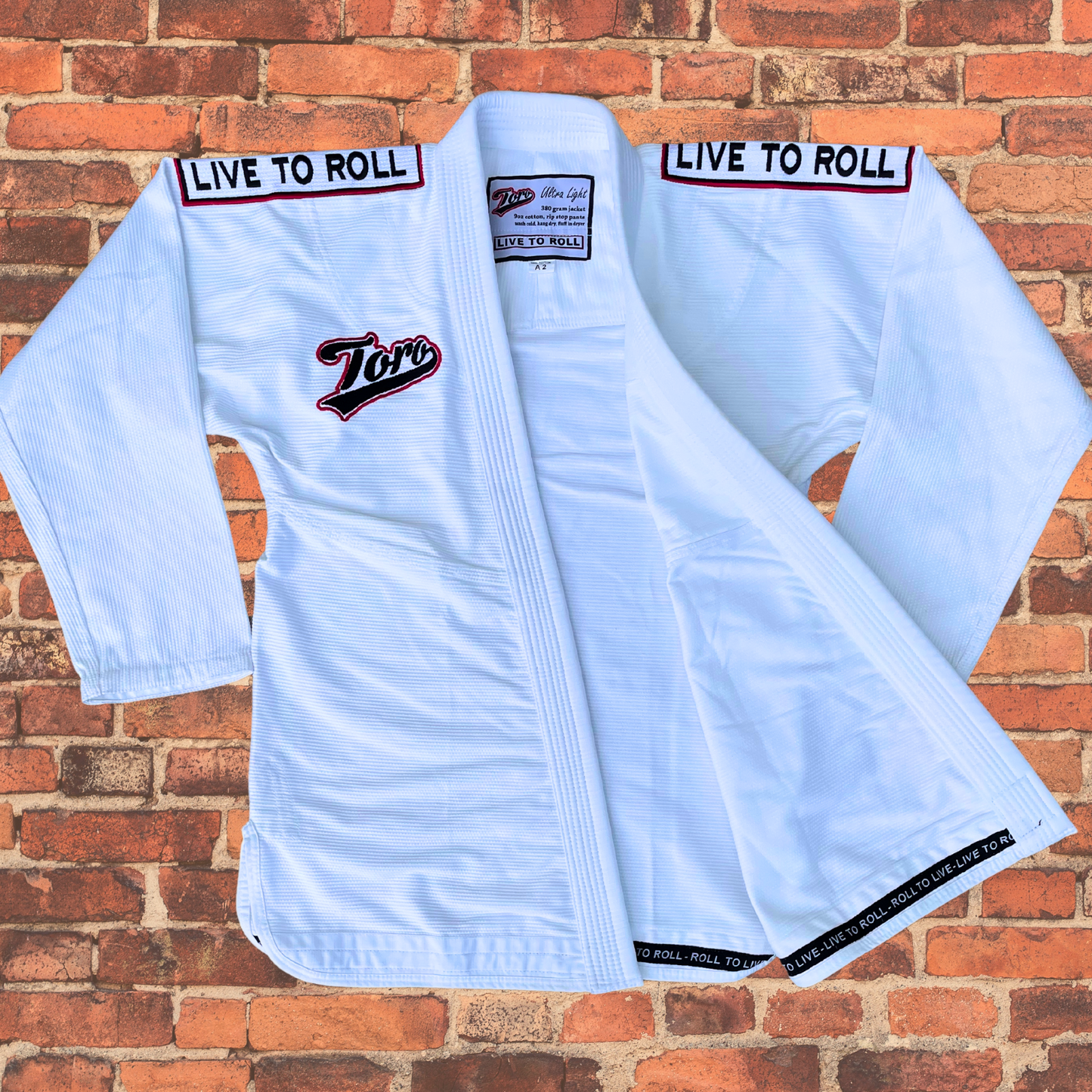 Close-up of Toro's white BJJ Gi jacket with 'Live to Roll' embroidered on the shoulders and Toro logo on the chest. The lightweight jacket is displayed open, revealing its durable and breathable fabric, ideal for Brazilian Jiu-Jitsu training and competition. Set against a red brick wall background.